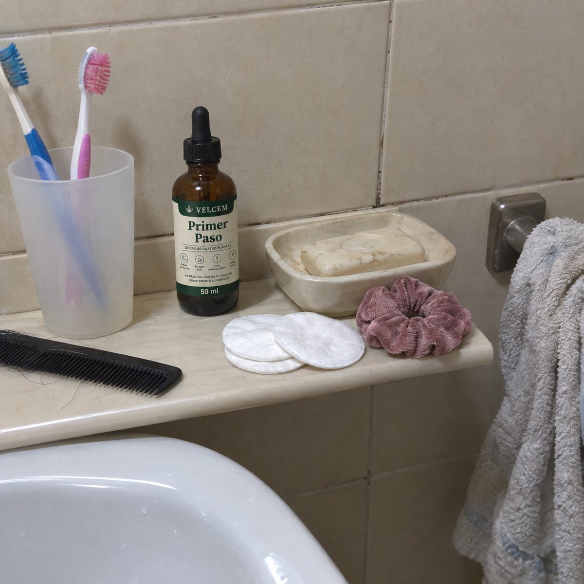 Foto de móvil de la repisa de un baño de azulejo beige: el frasco de VELCEM entre un vaso con dos cepillos de dientes, un peine, la jabonera, unos algodones y un coletero, junto a la toalla