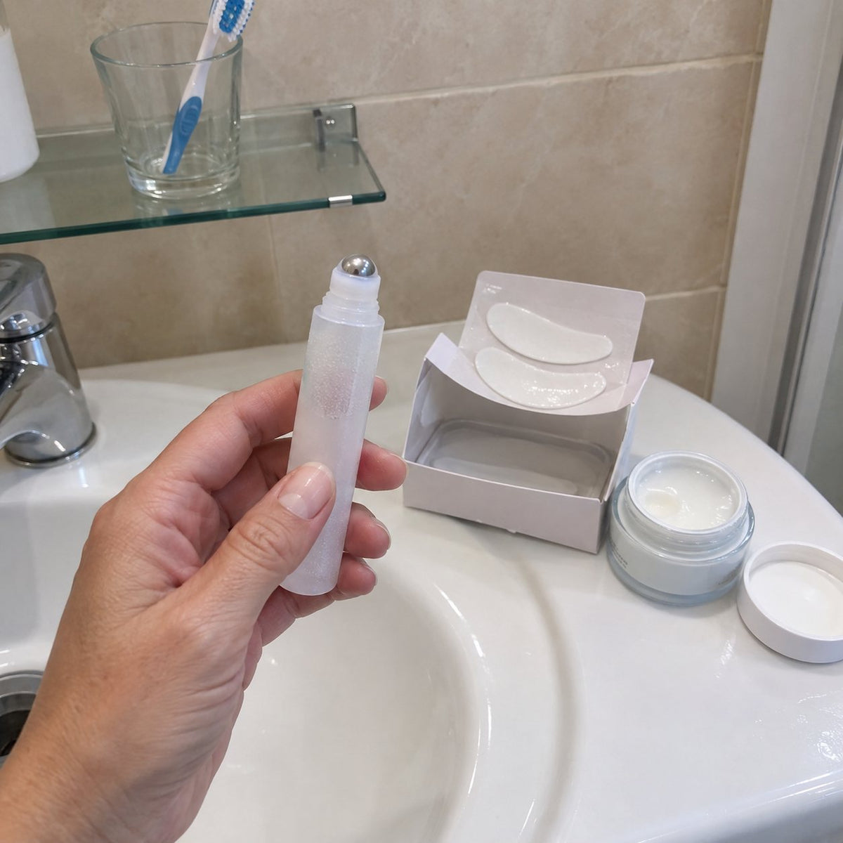 Su mano sostiene un contorno de ojos roll-on junto a una caja abierta de parches para los ojos y un tarro de crema abierto, en el borde del lavabo de su baño