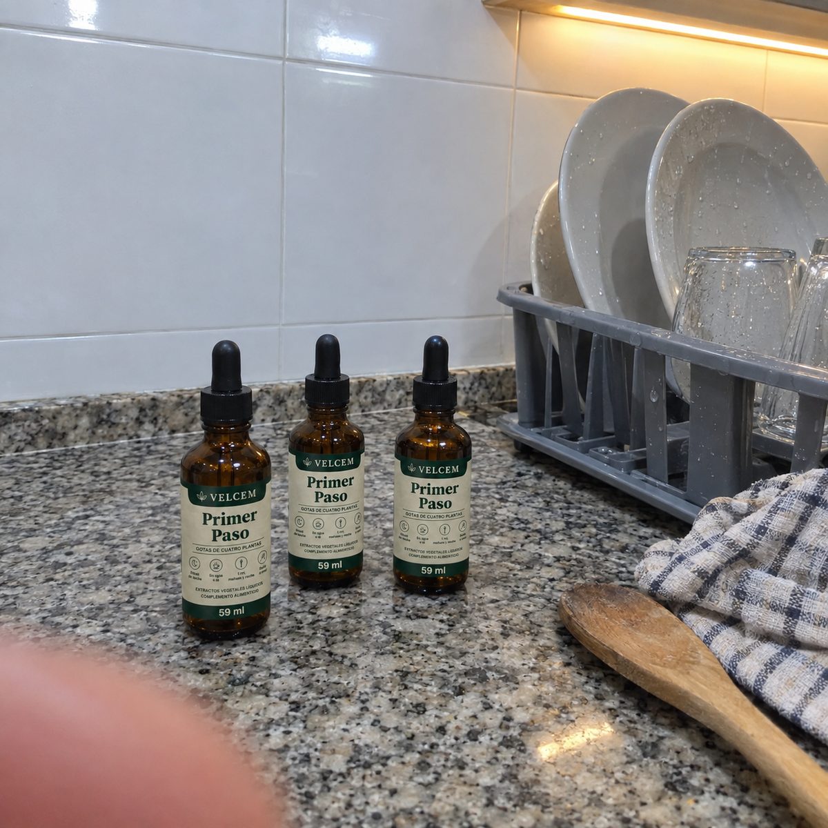 Foto de móvil de la encimera de granito de una cocina: tres frascos de VELCEM junto al escurreplatos con los platos fregados, un paño de cuadros y una cuchara de palo; un dedo tapa una esquina