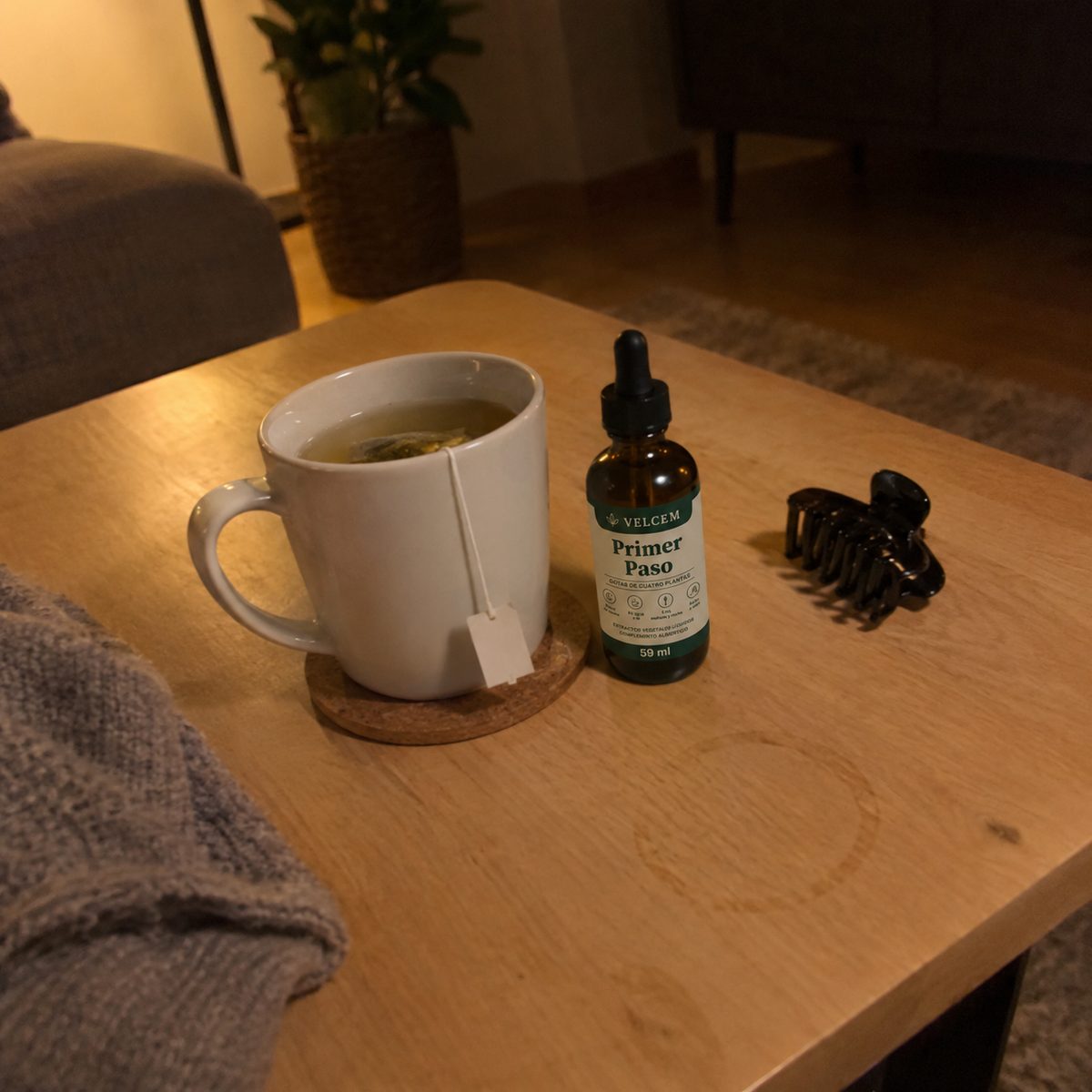Foto de móvil, con poca luz, de la mesa baja del salón: una taza de tila con su bolsita, el frasco de VELCEM y una pinza del pelo, junto al sofá y una manta