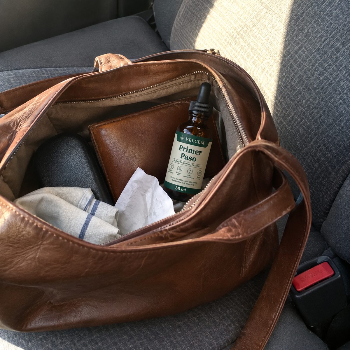 Foto de móvil de un bolso de piel marrón abierto en el asiento del coche, al sol de la tarde: el frasco de VELCEM asoma junto al monedero, unos pañuelos de papel y un pañuelo de tela